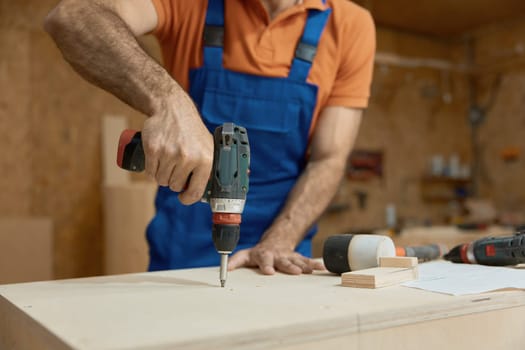 Cropped midsection shot with male carpenter working with drill electric screwdriver making holes in wooden plank. Woodworking and furniture production process