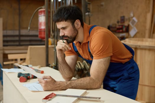 Pensive man carpenter reading blueprints analyzing and thinking about project during woodworking and furniture production process at workshop