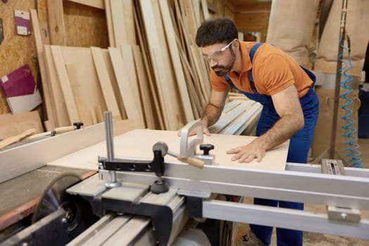 Young craftsman in uniform and protective goggles cutting woods on pieces working at circular cutting table in furniture factory workshop