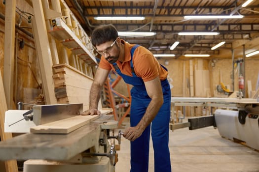 Man professional carpenter wearing workwear using jointer equipment for final processing of wood plank. Furniture production industry and woodworking concept