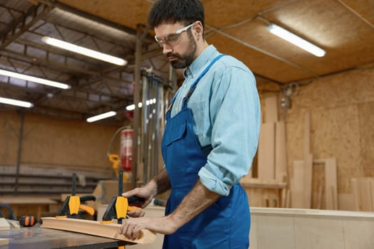 Carpenter fixing wooden plank on workbench with clamps carpentry tools while working at craft woodwork studio