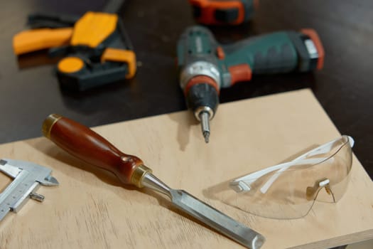 Carpentry manual tools and wooden board on workbench closeup. Chisel, ruler, electric drill power instrument, protective plastic goggles arrangement
