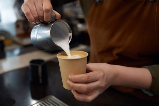 Closeup barista pouring steamed warm milk into disposable paper cup of aroma coffee drink over worktop