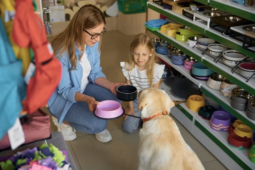 Mother and toddler daughter choosing new plastic food plate for Labrador dog enjoying shopping at pet store