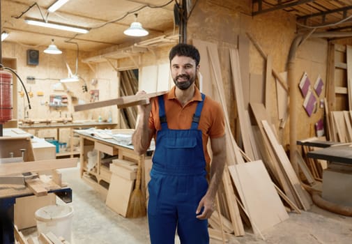 Smiling woodworker carrying timber plank on shoulder standing over carpentry workshop background. Woodworking and furniture manufacturing concept