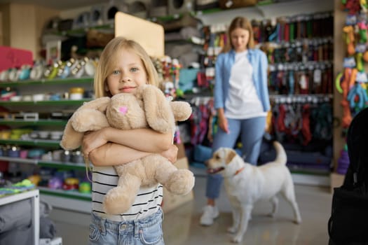 Little girl hugging soft bunny toy standing over pet store with mother and dog on blurred background