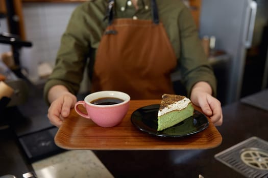 Cropped shot of woman barista giving wooden tray with freshly brewed coffee drink in ceramic cup and cake piece portion on plate. Client service at cafeteria or restaurant