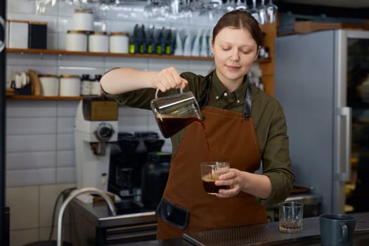 Woman barista pouring transparent brown aroma coffee drink in cup from glass jug. Process of high quality aromatic beverage preparation