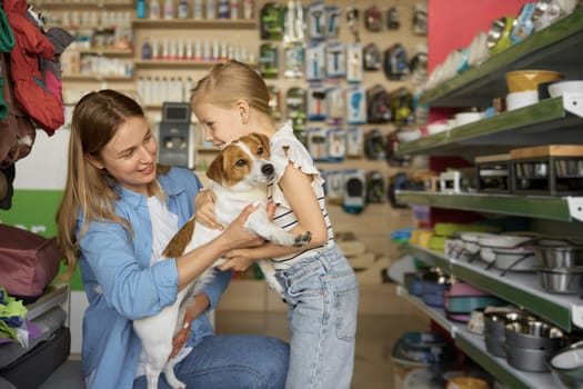 Mother helping cute little girl posing with dogs in hands over pet shop background. Happy time together and family shopping enjoyment