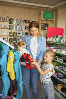 Happy mother and daughter buying new winter clothes for dog choosing red warm coat at pet shop rack