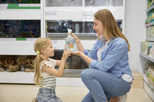 Happy satisfied mother and girl child holding new gold fish in plastic packet with water kneeling over pet shop background