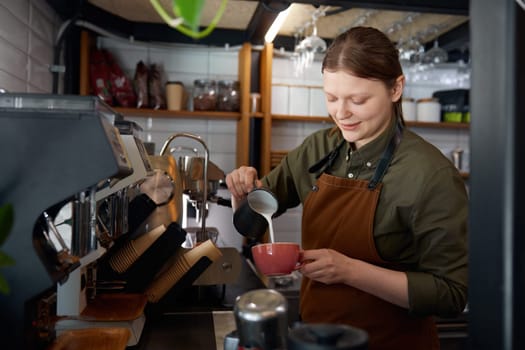 Young experienced woman barista in apron pouring milk into coffee cup with freshly brewed drink. Coffee-shop or cafe staff servicing clients