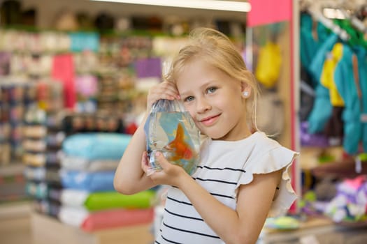 Portrait of cute smiling little girl holding packet with new gold fish pet standing over supermarket vet department store background