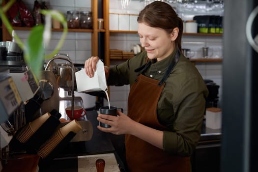 Young experienced woman barista in apron pouring milk into coffee cup with freshly brewed drink. Coffee-shop or cafe staff servicing clients