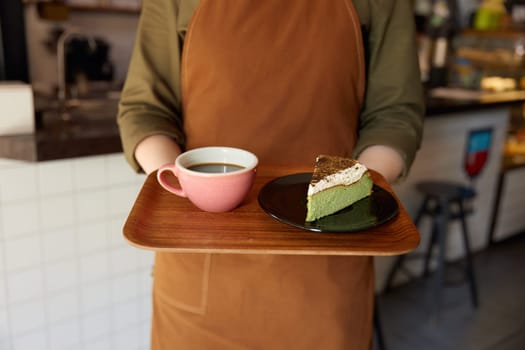 Cropped shot of woman barista giving wooden tray with freshly brewed coffee drink in ceramic cup and cake piece portion on plate. Client service at cafeteria or restaurant