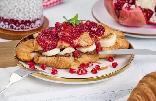 Croissant with banana pieces, chocolate paste and pomegranate seeds in a round white plate