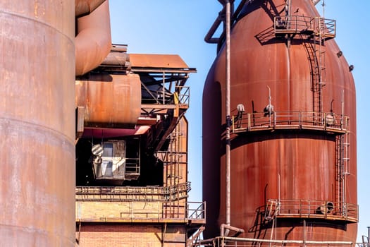 Abandoned blast furnace casting rusty hue over industrial landscape, contrasting with blue sky and fluffy clouds in scene of urban decay