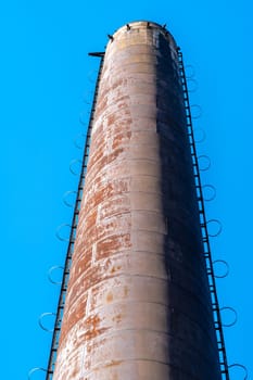 Tall rusty metal chimney with a ladder on the side stands out against a clear blue sky, showcasing industrial architecture