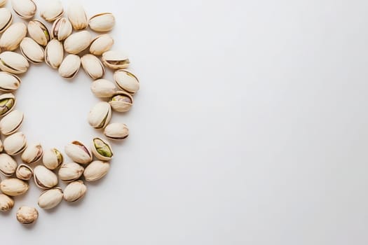 A group of pistachios is artistically arranged in a circular layout on a smooth white surface, highlighting their natural textures and colors. The simple composition draws attention to the nuts.