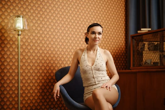 Relaxed woman sits elegantly in a comfortable chair against a background of vintage wallpaper next to a floor lamp