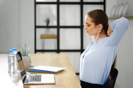A woman in a light blue shirt stretches and relaxes at her desk, taking a break from work on her laptop. Water bottles and a notebook are visible, emphasizing a focused yet mindful work environment