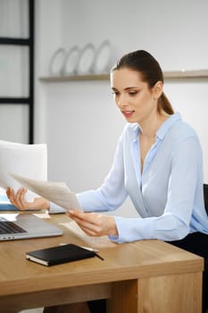 In a contemporary office, a business woman sits at her desk, closely examining paperwork while using a laptop. She appears focused and engaged, highlighting the importance of her tasks.