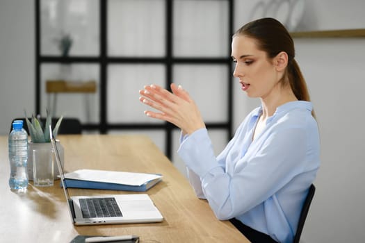 A woman in a light blue blouse participates in a video call, using a laptop placed on a wooden desk.