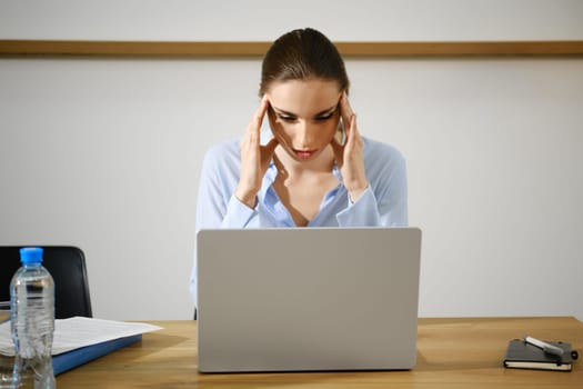 A woman sits at a desk in a contemporary office, visibly stressed as she rubs her temples while looking at a laptop screen.