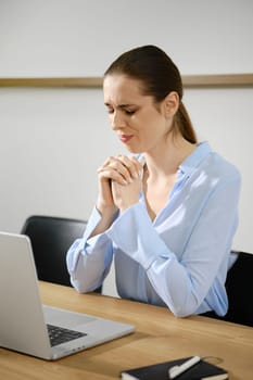 A woman wearing a blue shirt is seated at a wooden table, looking pensive while praying with her hands clasped.