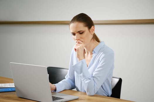 A woman sits at a wooden desk, thoughtfully working on her laptop. She appears focused and contemplative in a well-lit modern office setting, creating an atmosphere of productivity and concentration.
