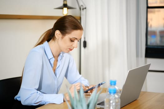 A woman in a light blue shirt is focused on her smartphone while seated at a wooden desk with a laptop. Natural light streams through the window, enhancing the contemporary office atmosphere.