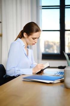 A woman in a light blue blouse sits at a wooden desk, focused on writing in a notebook. Sunlight pours through the nearby window, illuminating her workspace filled with a laptop and stationery.