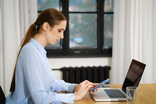 A woman sits at a wooden desk, focused on her smartphone while her laptop is open beside her. Natural light streams through the large windows, creating a calm workspace atmosphere.