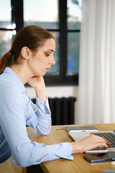 A woman in a light blue blouse is focused on her laptop, seated at a wooden desk in a well-lit office. Natural light streams in through large windows, enhancing the productive atmosphere.