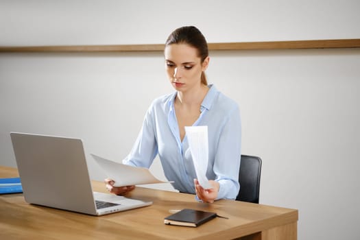 A woman is attentively examining important documents at a wooden desk, with a laptop open in front of her.