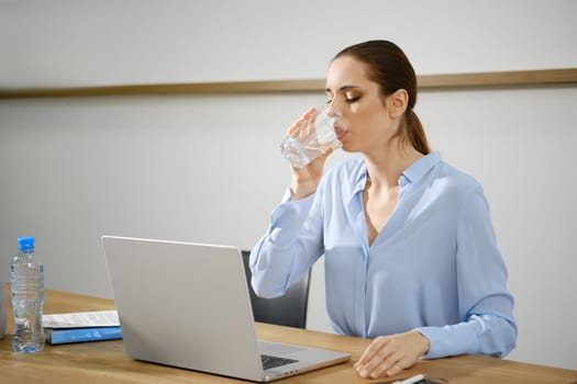 A woman in a light blue blouse pauses to sip water from a glass while working on her laptop at a wooden desk in a modern office environment, maintaining focus and hydration during her tasks