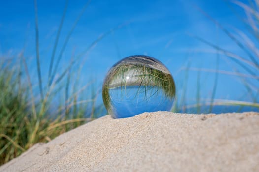 A glass ball lies in the sand of a dune by the sea, the dune grass and the blue sky are reflected in the ball