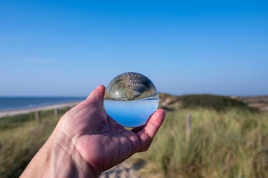 A human hand holds a glass ball, in the ball a sand dune, a footpath, blue sky and the sea are reflected