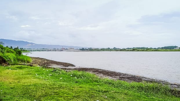 A river with a bridge in the background. The water is calm and the grass is green. The scene is peaceful and serene