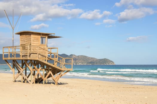 A wooden lifeguard tower on a sandy beach with ocean waves and mountains in view, mallorca balearic islands, spain