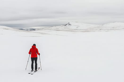 A lone skier glides across a vast expanse of untouched snow, surrounded by gentle hills under a soft gray sky. The quiet and stillness of the snowy terrain creates a peaceful atmosphere.