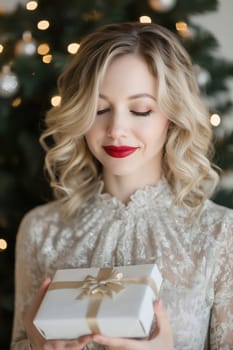 A woman with blonde curls smiles gently, holding a wrapped gift in front of a beautifully lit Christmas tree.