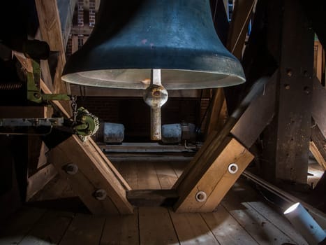 Heavy bell with clapper and ringing mechanism in the wooden church tower illuminated from the side by a light.