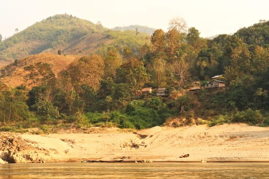View from the slow boat from Thailand to Laos onto the shore of the Mekong River with a small village, settlement with bamboo huts, cabins, Laos 2023