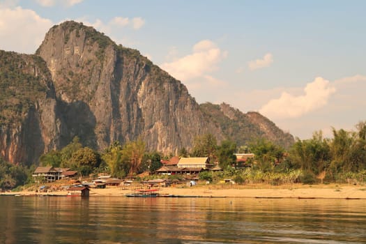 View from the slow boat from Thailand to Laos onto the shore of the Mekong River, a traditional village is reflecting in the water in front of a huge karst cliff, rock, Laos 2023