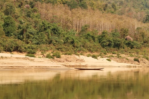 View from the slow boat from Thailand to Laos onto the shore of the Mekong River, a docked fisher boat is reflecting in the water in front of a dense forest, Laos 2023