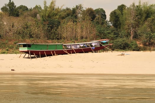View from the slow boat from Thailand to Laos, another traditional slow boat is jacked up on the beach, shore of the Mekong River, Laos 2023