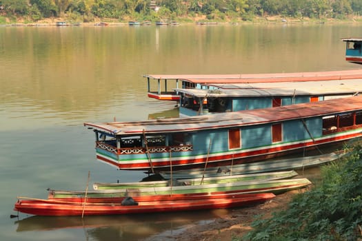 A few colorful long boats are docked on the shore of the Mekong River in the old town of Luang Prabang, Laos 2023