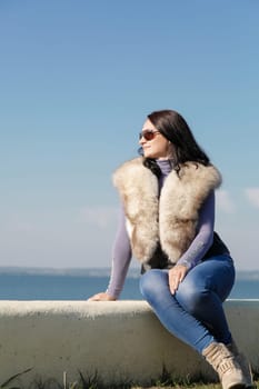 A stylish and fashionable woman in a fur vest sits on a pier against the background of the sea