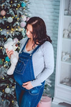 A pregnant woman is in a cozy setting, gently holding a teddy bear close to a beautifully festive tree nearby